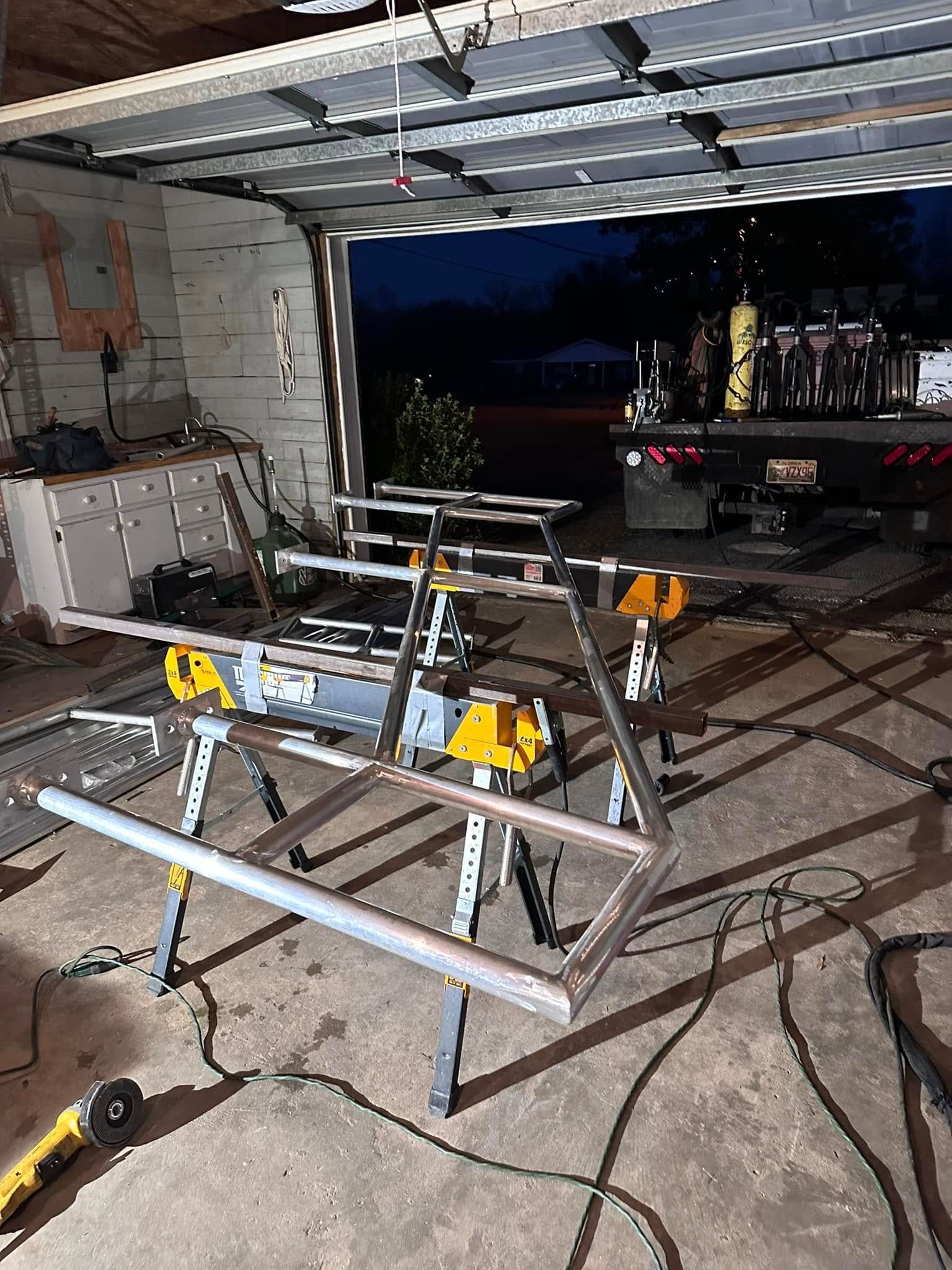 Welded steel frame on sawhorses inside a garage, with an open garage door showing the outside.