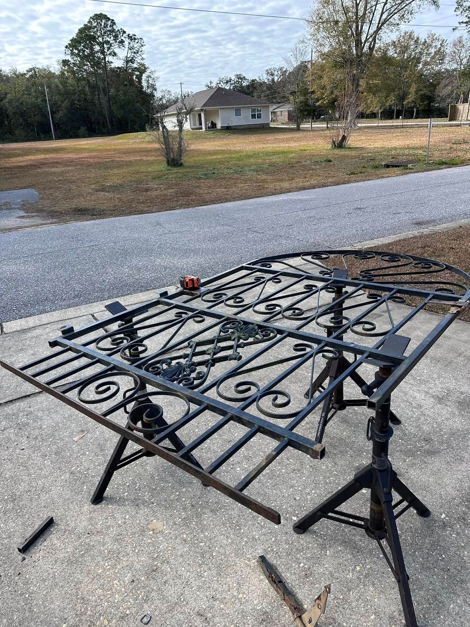 Black metal gate on sawhorses outdoors; gravel driveway and houses in background.