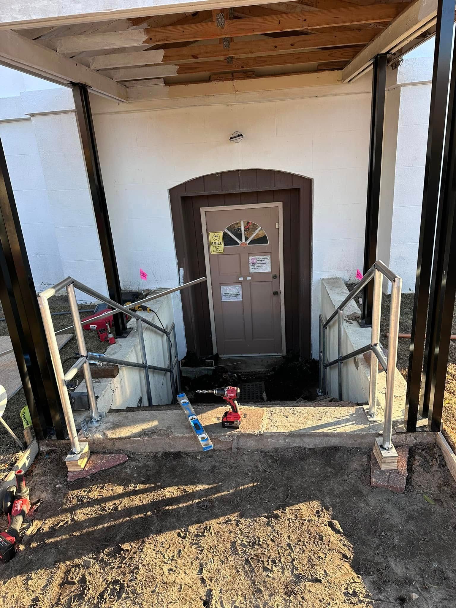 Exterior doorway with stairs, ramp rails. Construction, repairs happening. Brown door, white building, brown dirt.