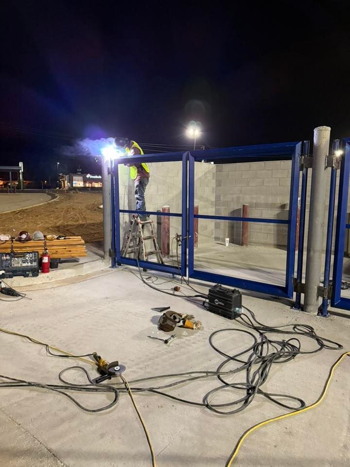 A worker welds on a blue metal gate at night. The setting is an unfinished concrete area.