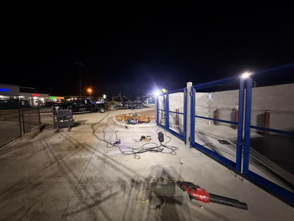 Nighttime construction site with blue gate. Tools, cables, and vehicles are visible.
