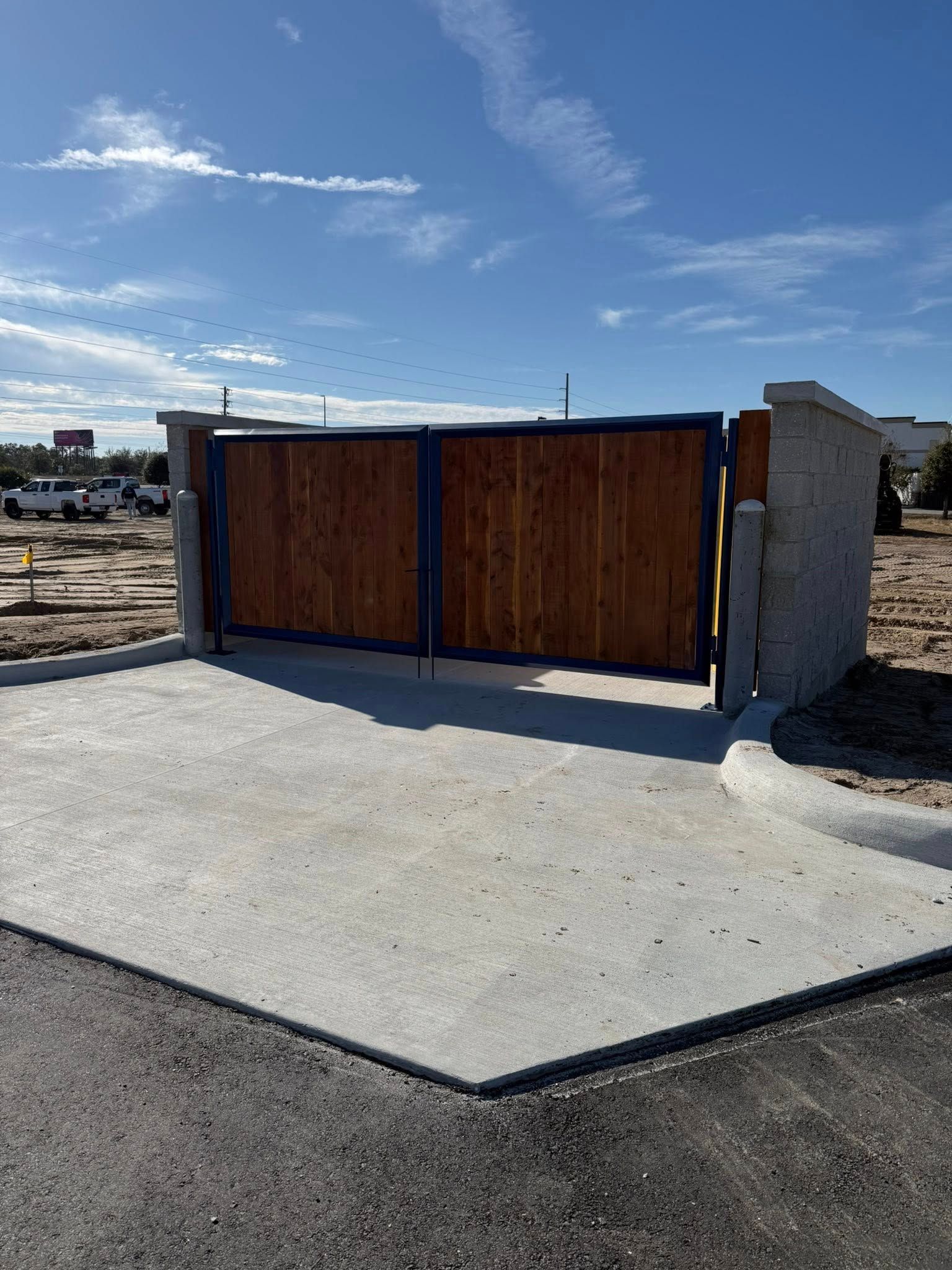 Wooden gate with blue frame, set in concrete, leading into a vacant lot under a blue sky.