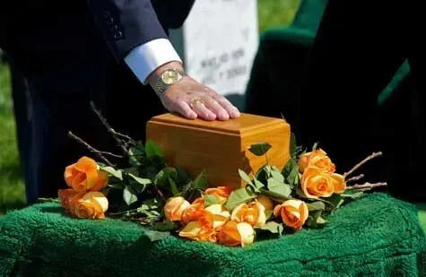 Person's hand on a wooden urn at a memorial. Orange roses and greenery rest on a green cloth.