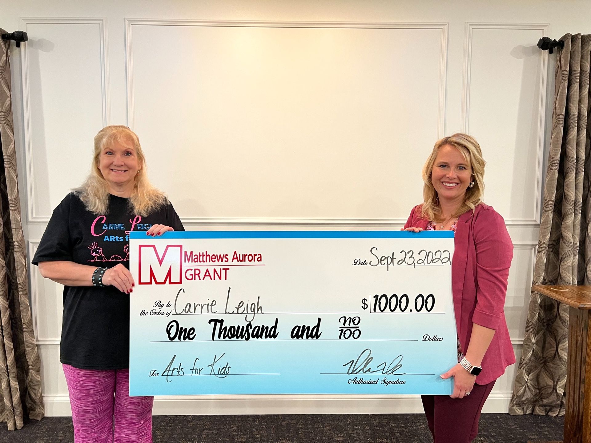Two women holding a large check, smiling, indoors. Check reads, 