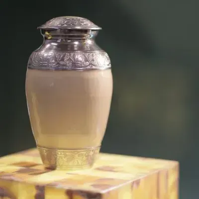 Cream-colored urn with silver trim and lid, on a marble-like pedestal, against a blurred green backdrop.