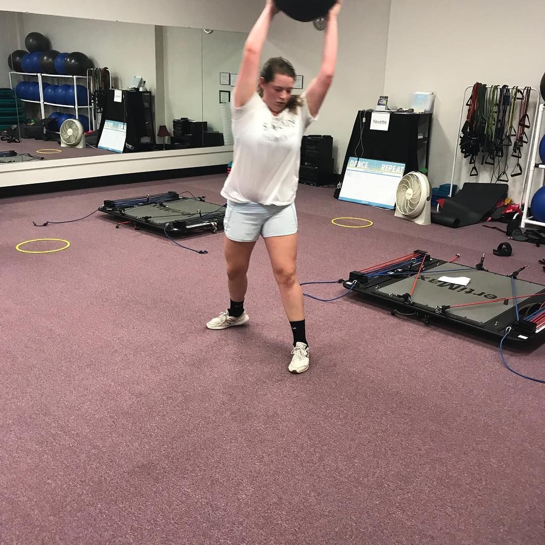 Woman in a gym, holding a medicine ball overhead, about to slam it down. She wears shorts and a white t-shirt.