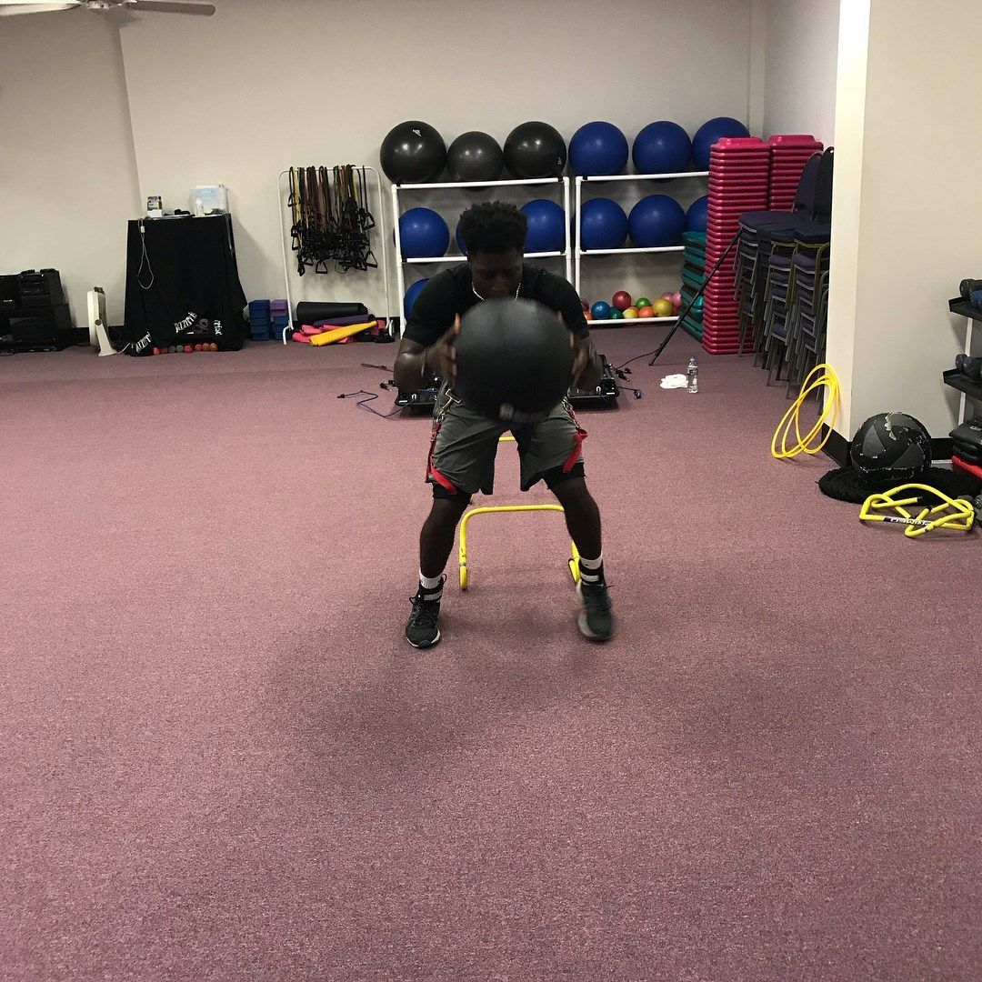 A person in a gym is holding a medicine ball, standing over a small step. The person wears shorts and athletic shoes.