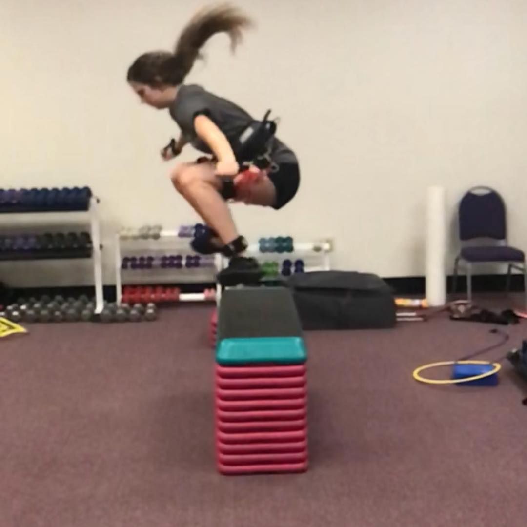 Woman jumps over a step platform. She is in athletic attire in a gym setting.