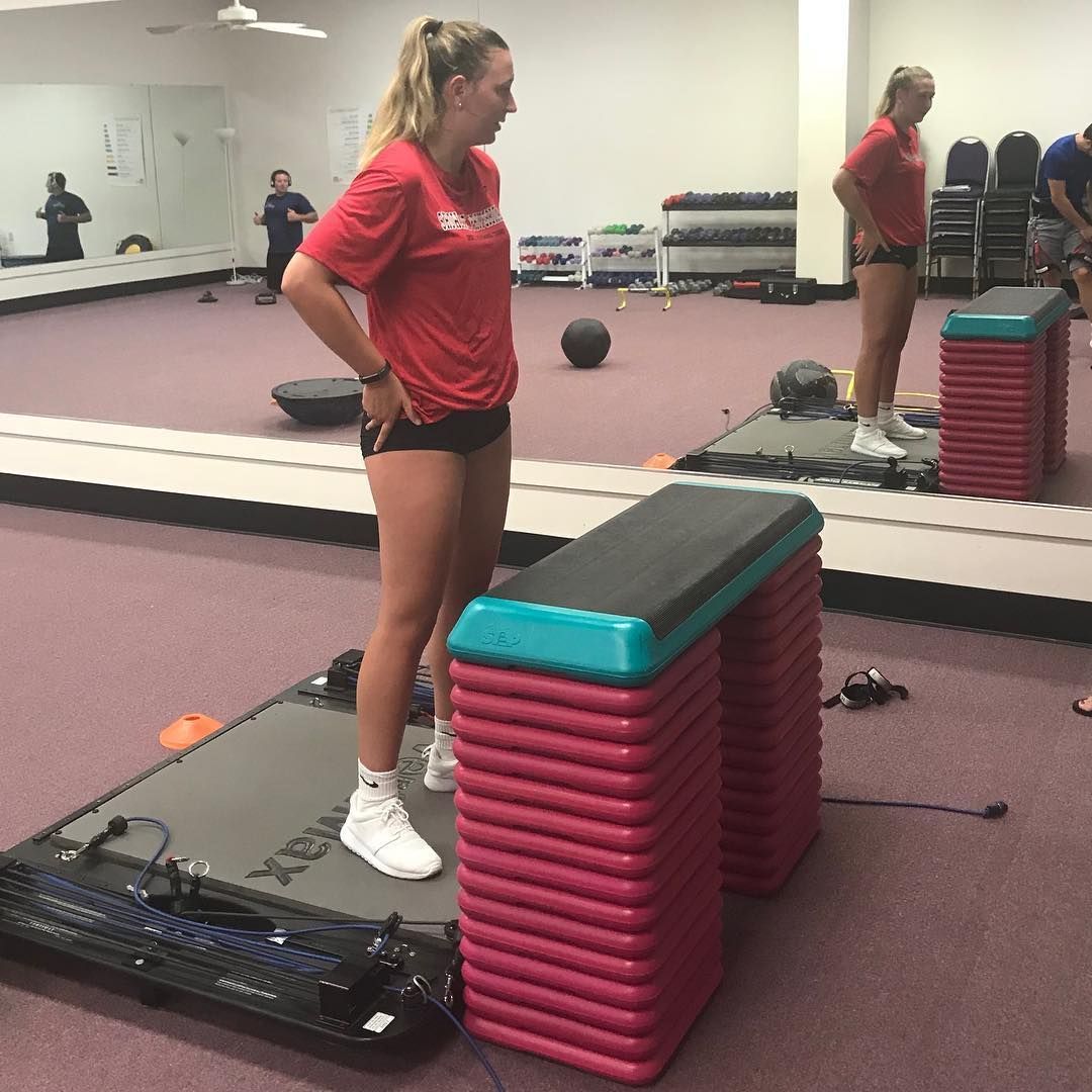 Woman in red shirt stands on a platform in a gym, facing a mirror, hands on hips, ready to exercise.