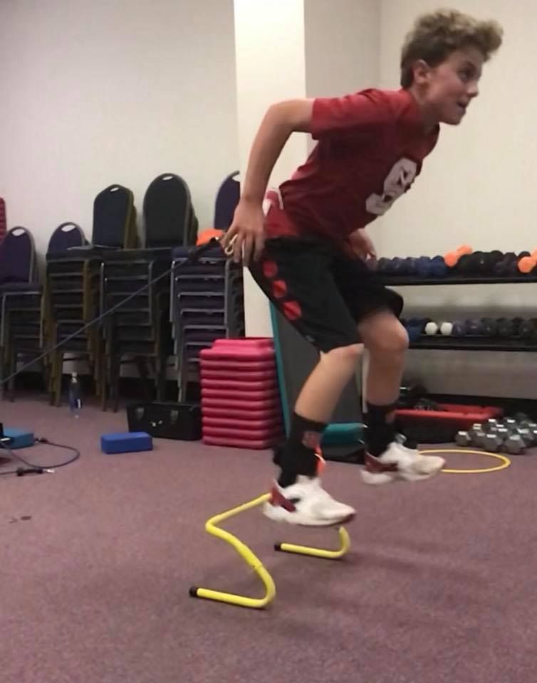 A young person jumps over a low yellow hurdle in a gym. They wear a red shirt, black shorts, and white sneakers.