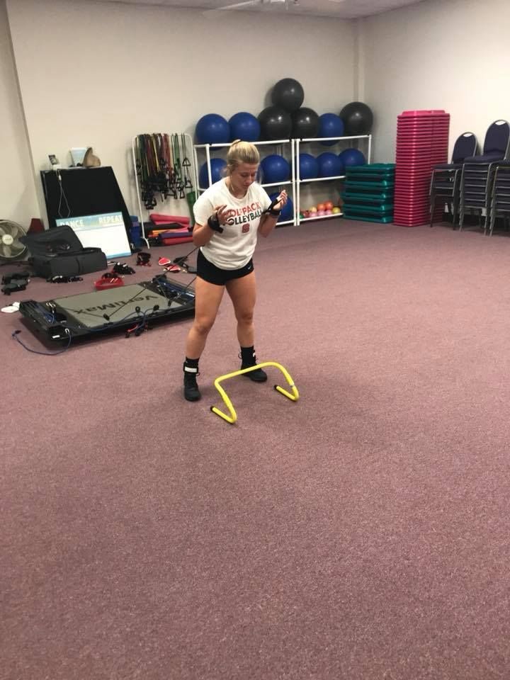 Woman exercising in a gym, straddling a low yellow hurdle, with other fitness equipment in the background.