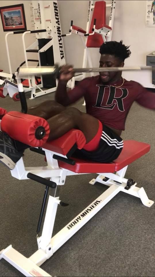 A young Black man does an ab workout on a machine, holding a barbell. He wears a maroon top, black shorts, and is in a gym setting.