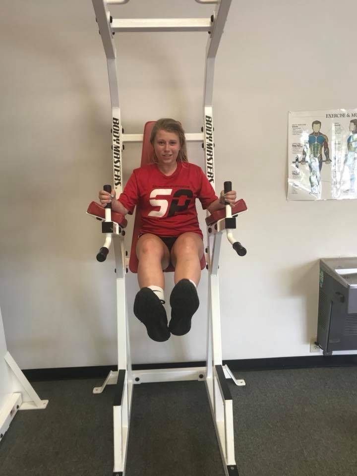 Woman in a red shirt doing a leg raise exercise on a vertical abdominal machine in a gym.