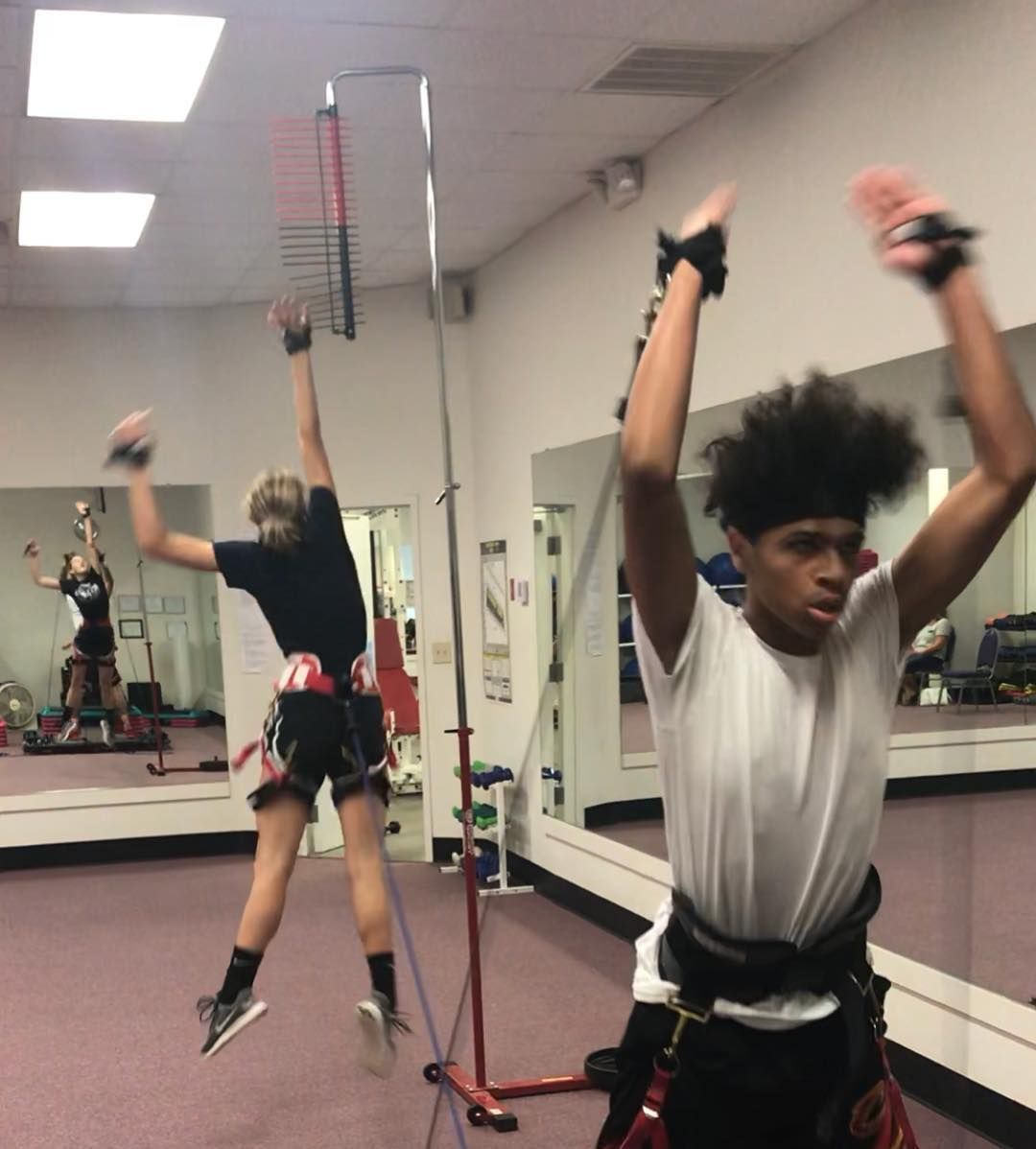 Two young men exercising, using bungee cords in a mirrored gym. They are jumping, arms raised.