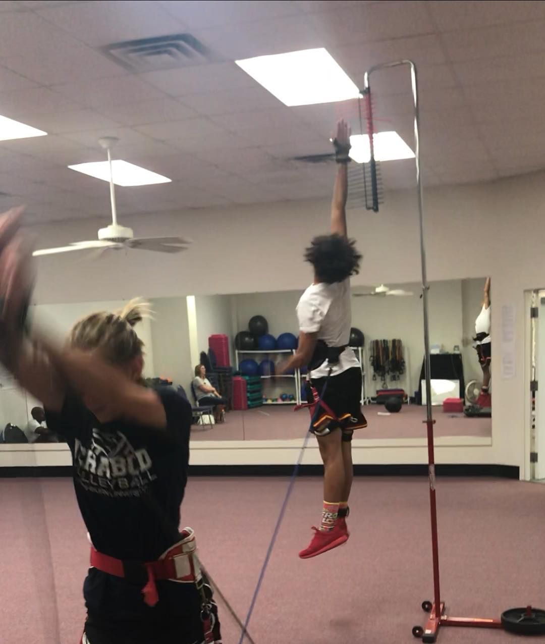 A person in a harness leaps upwards, connected to a high rod and rope, while another person watches in a gym setting.