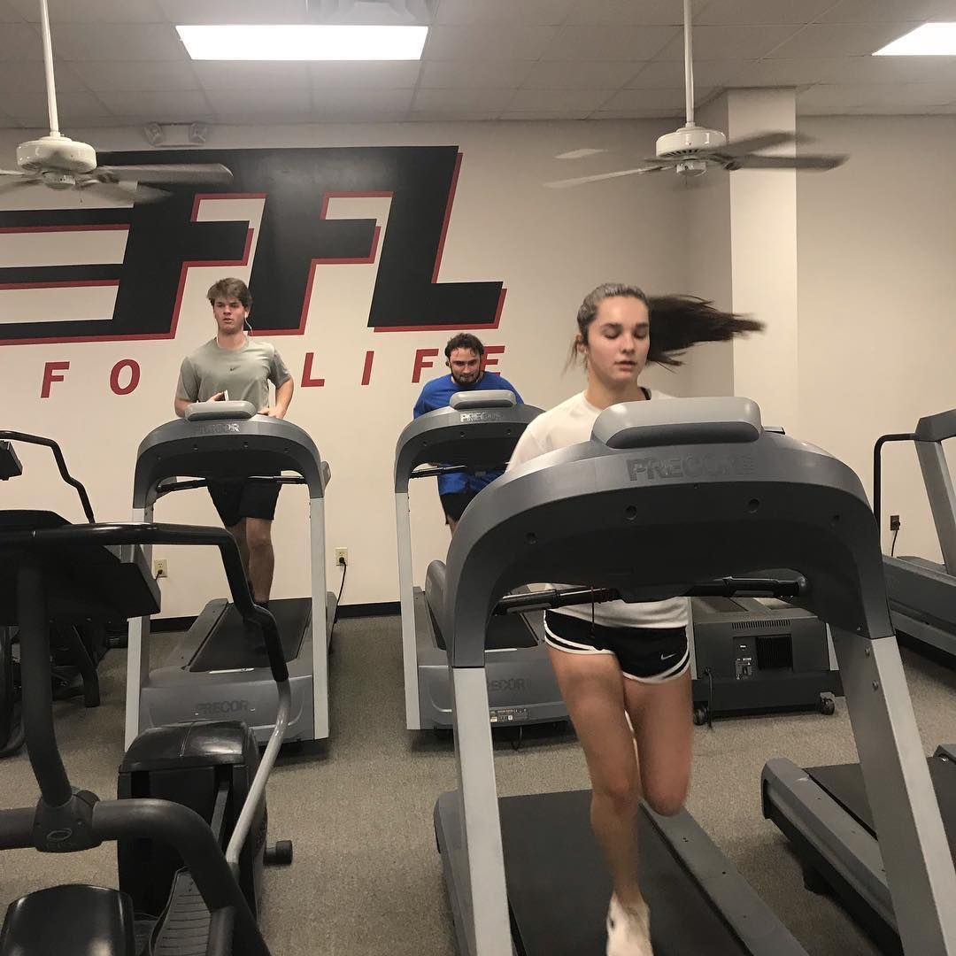 Three people running on treadmills in a gym. A woman in shorts runs with her hair flying back. The sign reads 