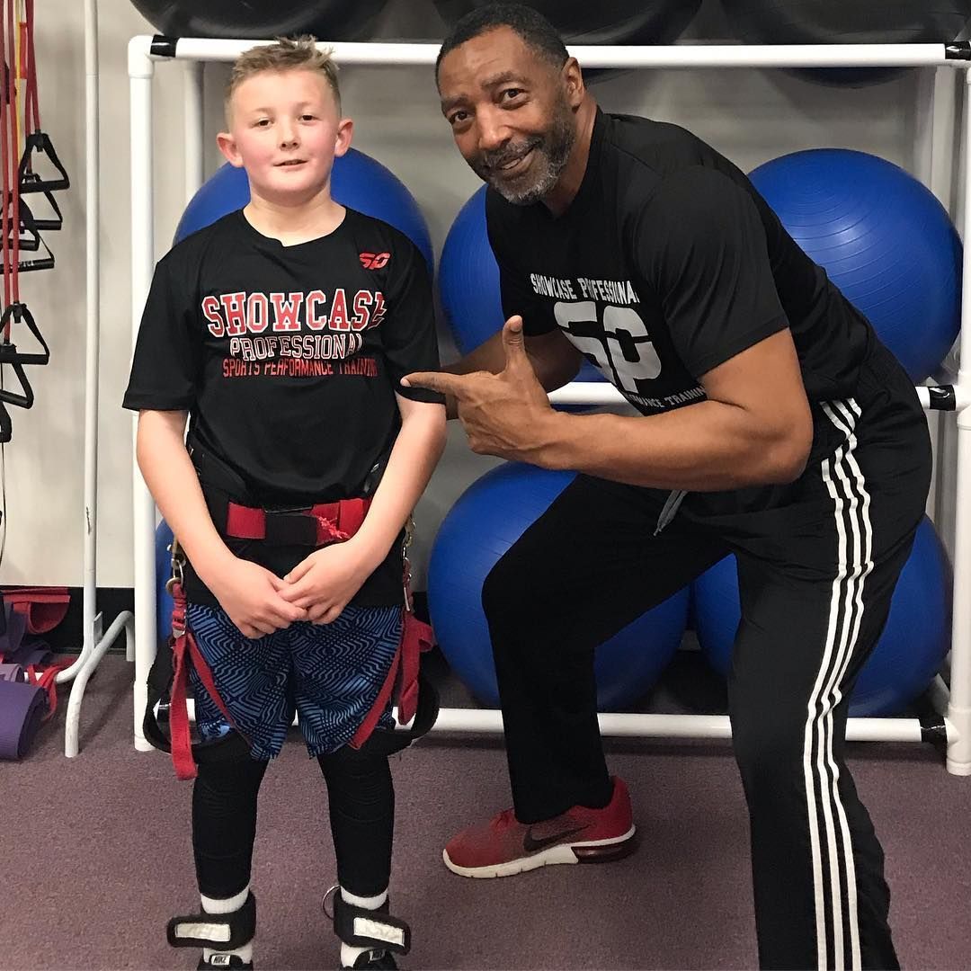 A young boy in workout gear stands next to a smiling Black man. Both are in a gym, near exercise balls and equipment. The man is pointing at the boy.