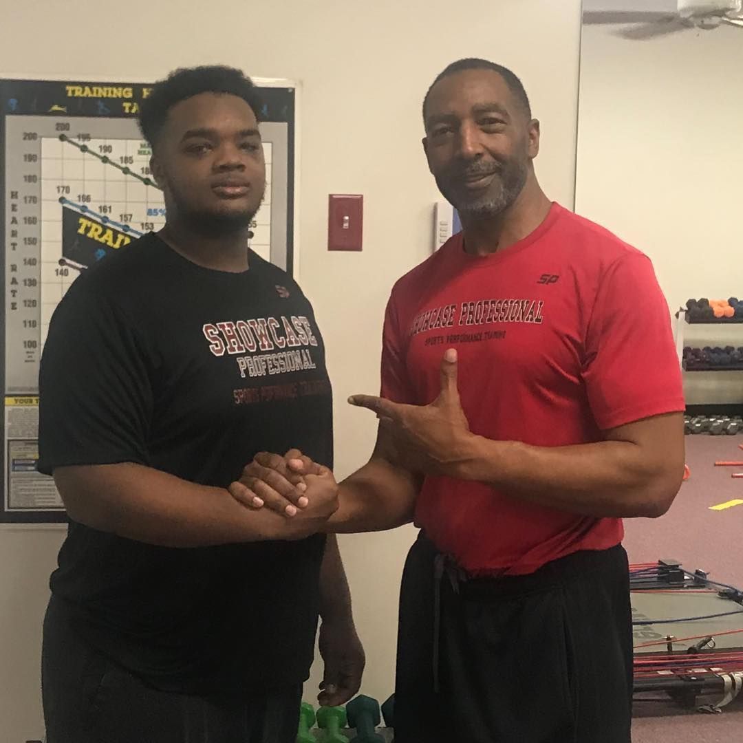 Two men in a gym shake hands. One, wearing black, smiles. The other, in red, points and smiles, with workout equipment visible behind them.