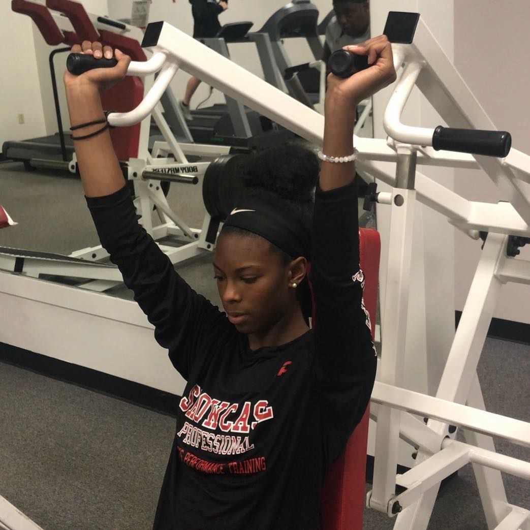 A person in a black long-sleeve shirt using a shoulder press machine in a gym, arms extended upwards.