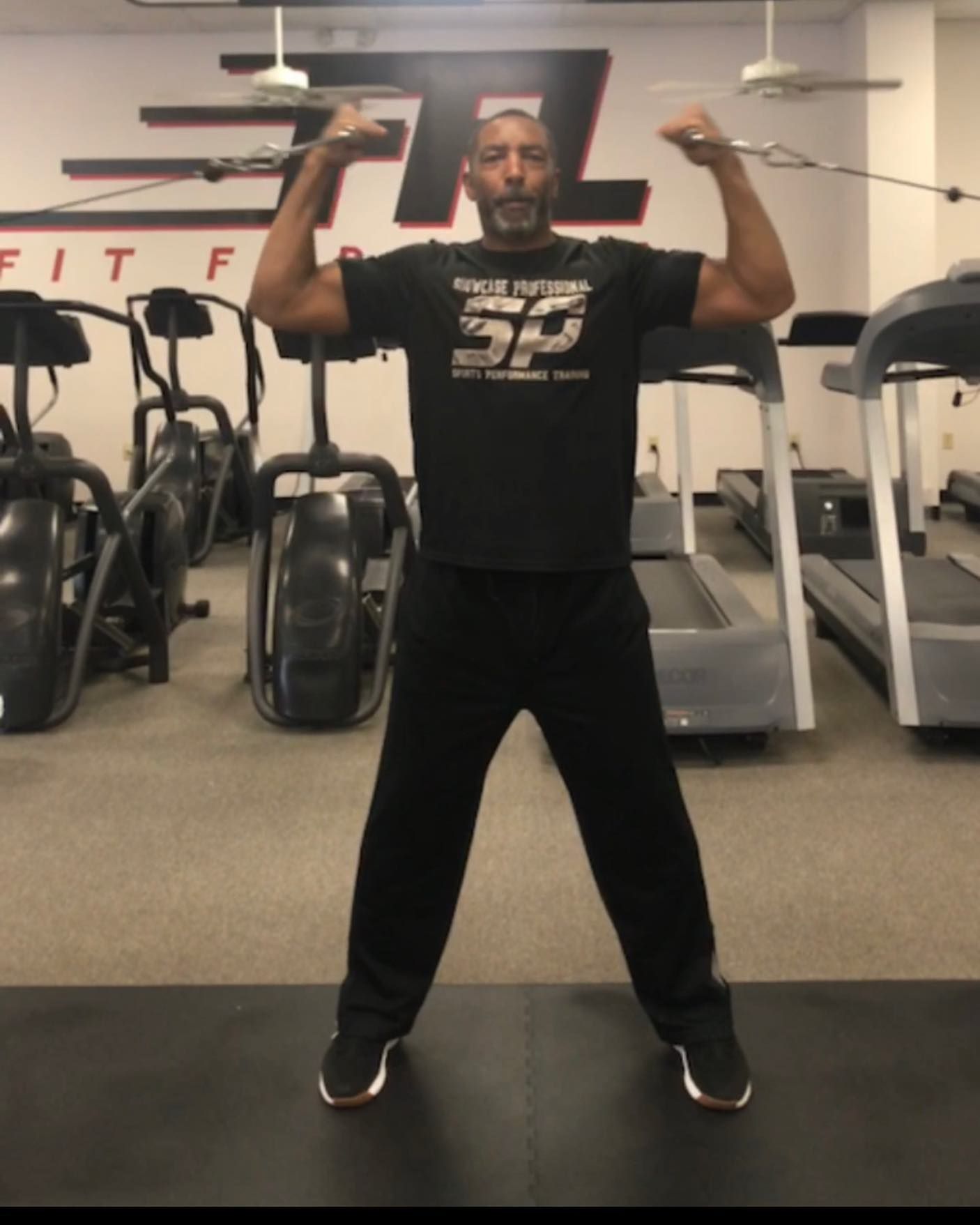 Man in black sweatsuit flexing biceps in a gym, with exercise equipment in the background.