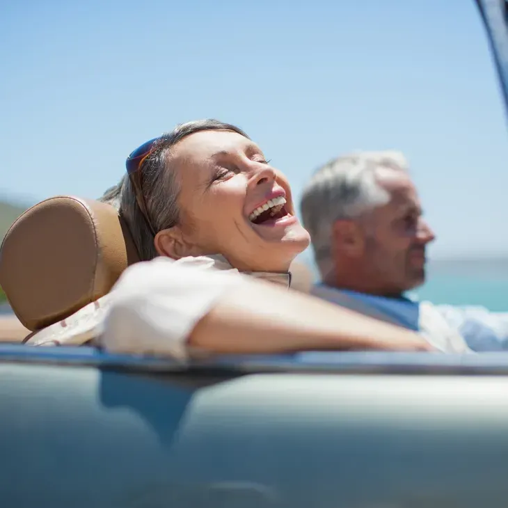 Una persona sonriente, recostada con los ojos cerrados en un descapotable en un día soleado, con un pasajero al fondo.