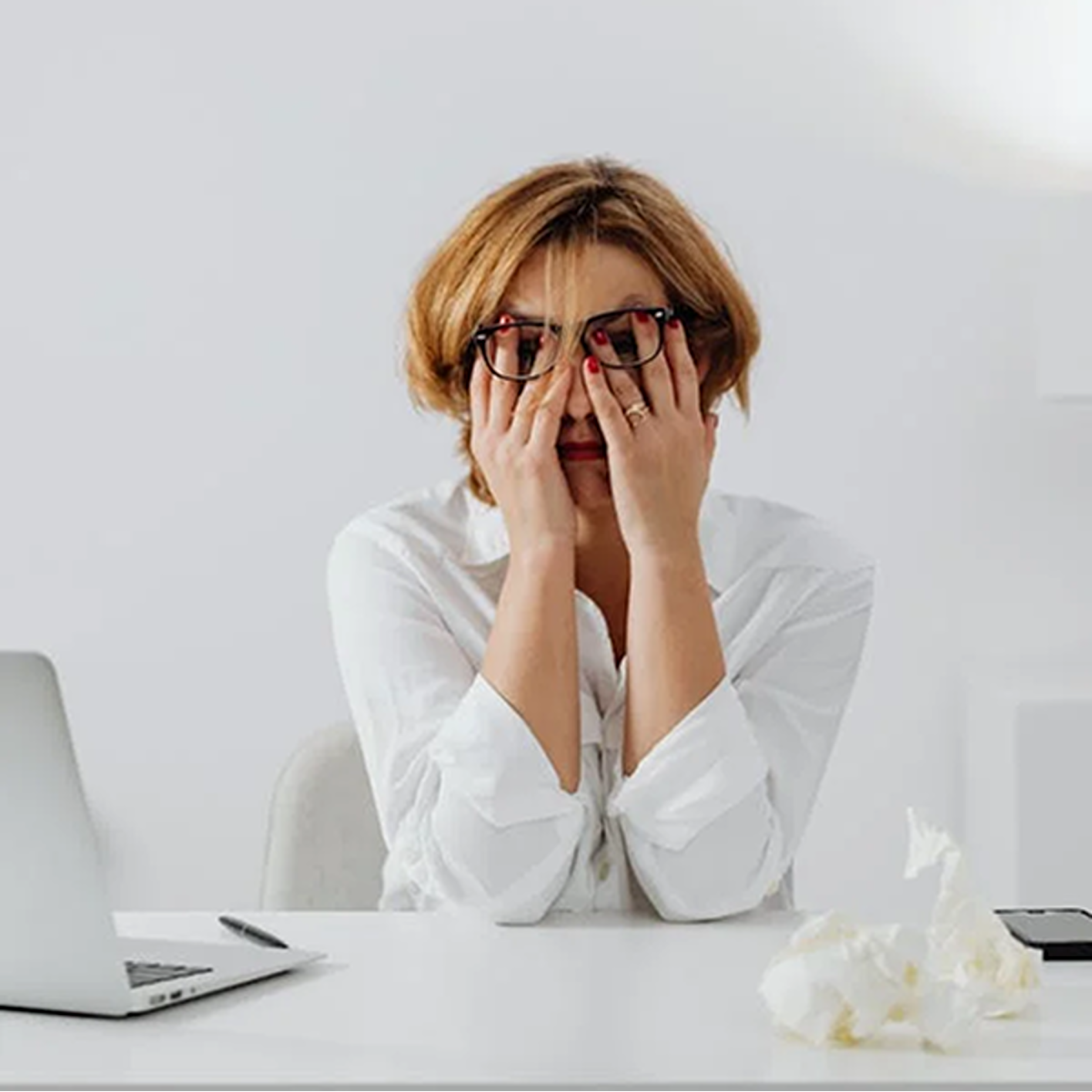 A woman wearing glasses is sitting at a desk with her hands on her face.
