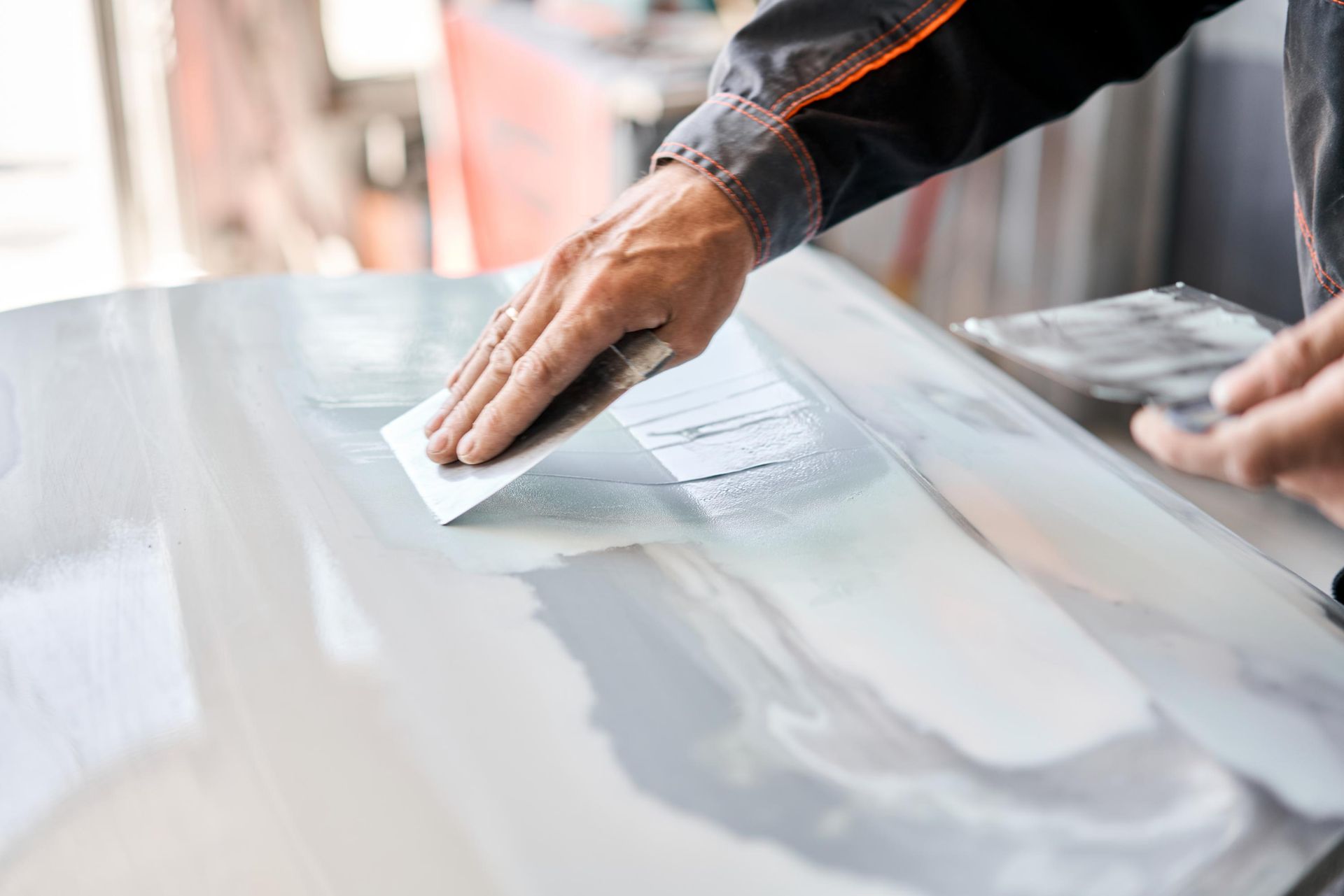 Person smoothing body filler on a car panel with a putty knife in a repair shop.