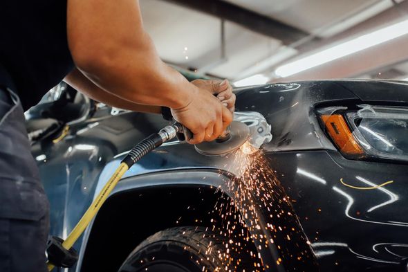 Person uses a grinder on a car fender, sparks flying in an auto repair shop.