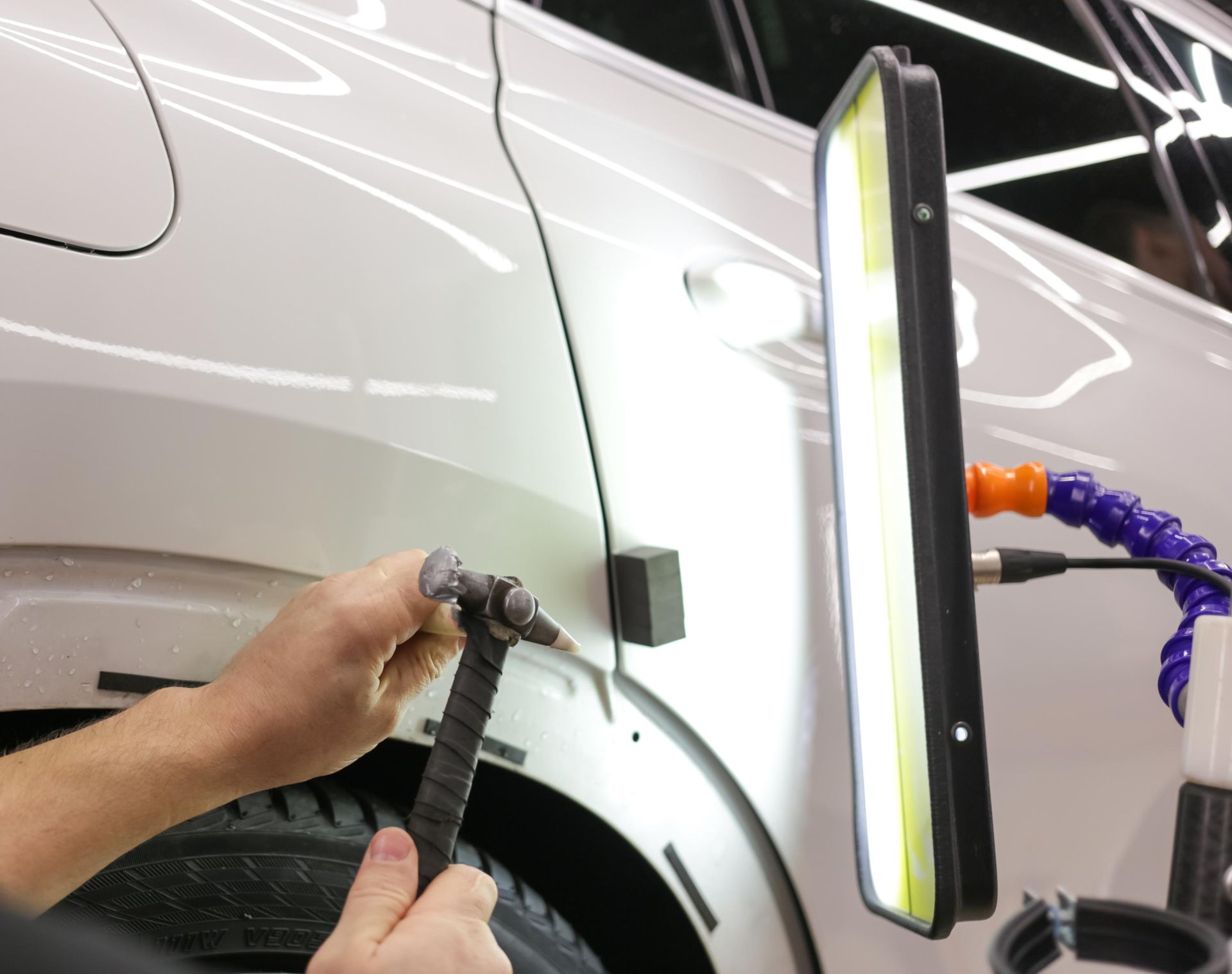 Person using tools to remove a dent from a white car's panel, illuminated by a specialized light.