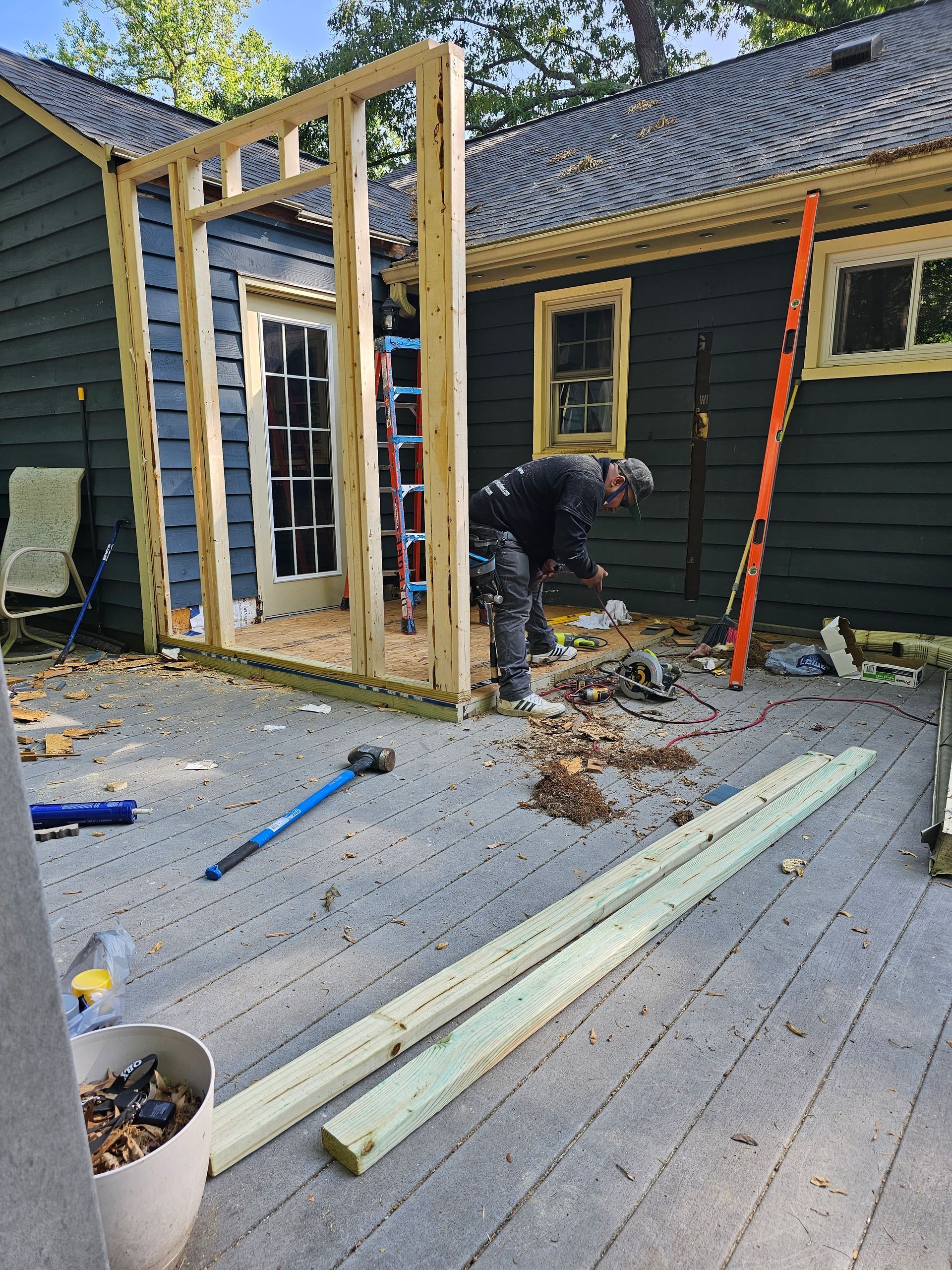 Workers framing a new doorway on a house deck, with tools and lumber nearby.