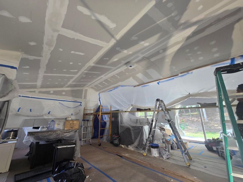 Interior of a room under renovation, with ladders, tools, and plastic sheeting covering walls and windows.