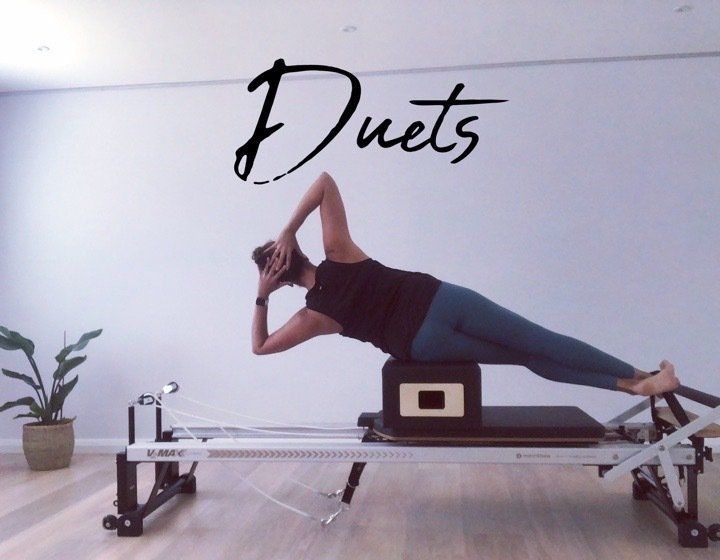 A woman is doing a pilates exercise in front of a wall that says duets