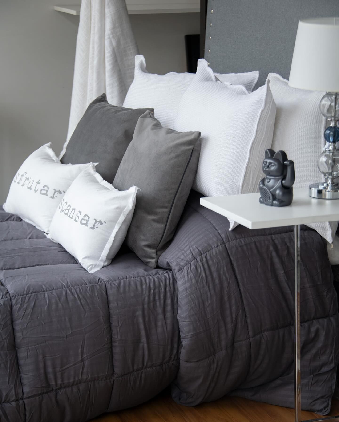 Cama con edredón y almohadas grises, mesita de noche blanca con decoración.