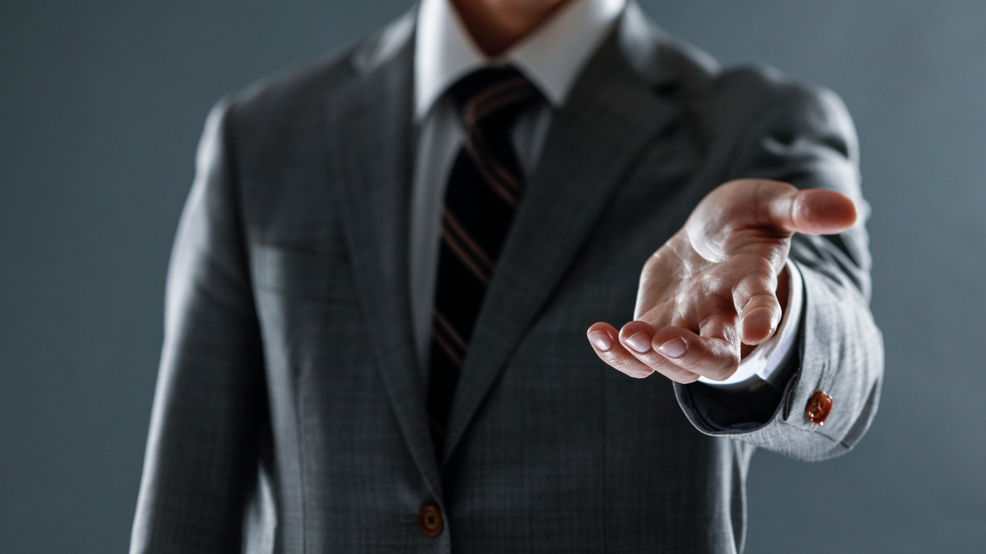 Man in gray suit extends hand, palm up, suggesting offer or request.