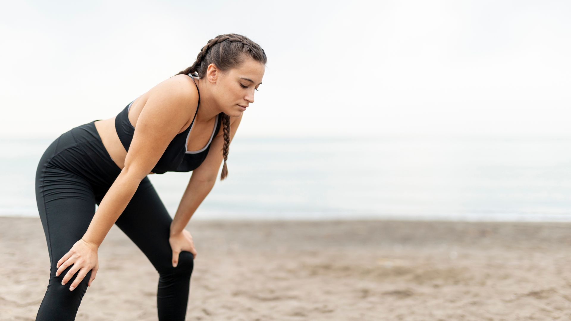 Mujer en ropa deportiva, inclinada, con las manos sobre las rodillas, en una playa.