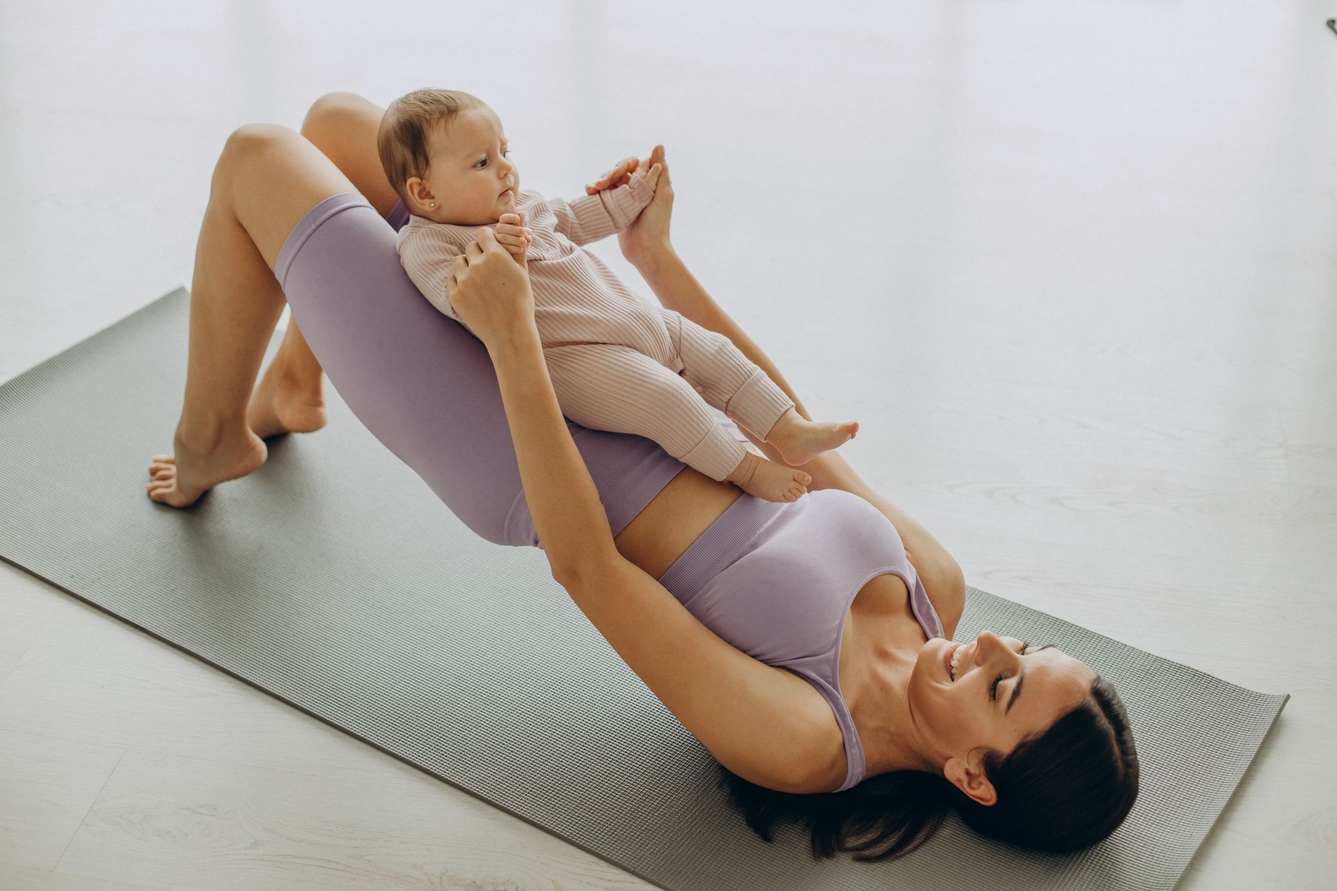 Mujer haciendo ejercicio sobre una colchoneta con un bebé en las caderas; en interiores, ropa deportiva de color violeta claro.