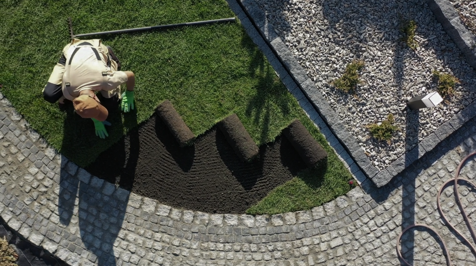 Person installing sod rolls in a yard, near a stone pathway and gravel area.