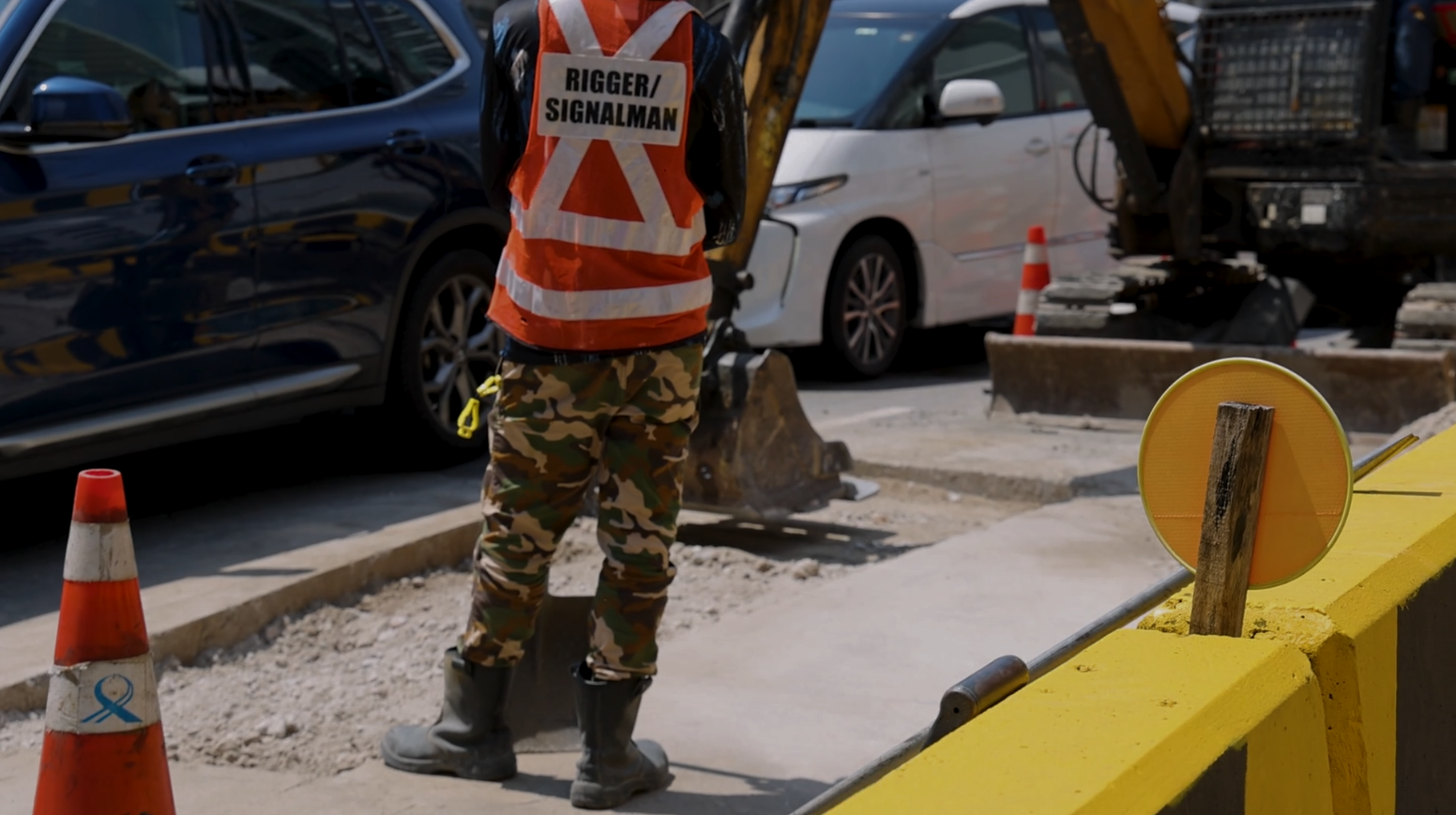 Construction worker directing traffic near a road excavation. Orange vest, camouflage pants, vehicles and equipment visible.