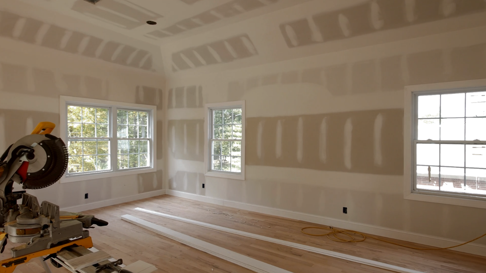 Room under construction with drywall installed. Windows, miter saw, and wooden floor visible.
