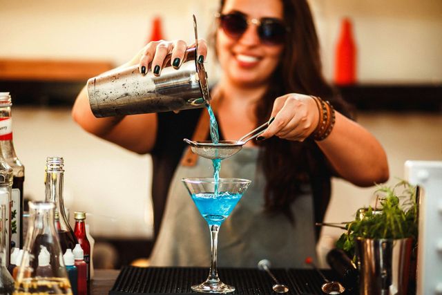 Bartender smiles, straining blue cocktail into a martini glass. Black shirt, sunglasses, outdoor setting.