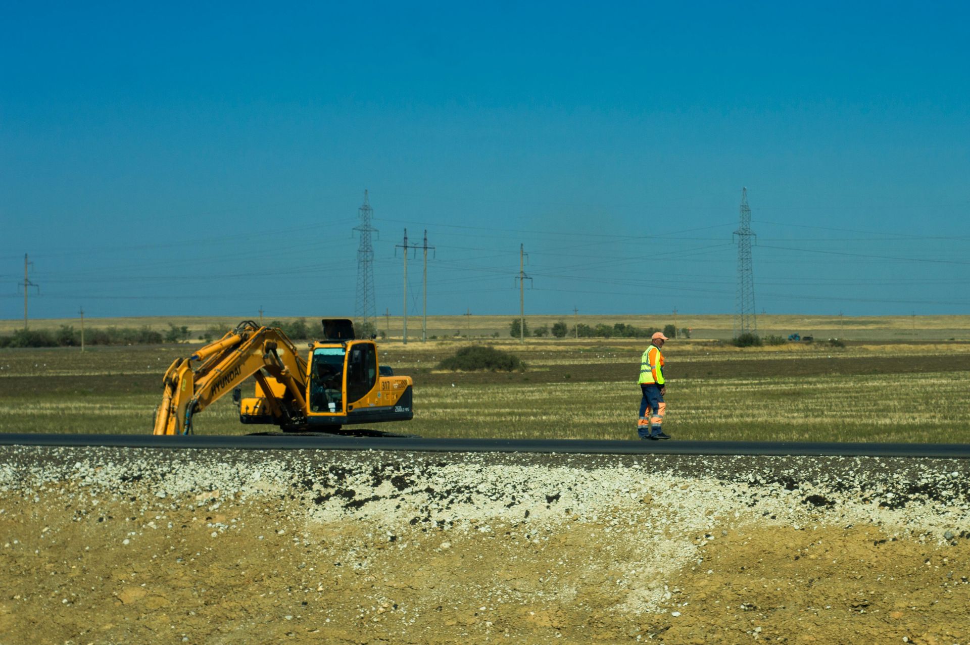 Yellow excavator and worker in reflective vest on a road, with a blue sky and fields in the background.