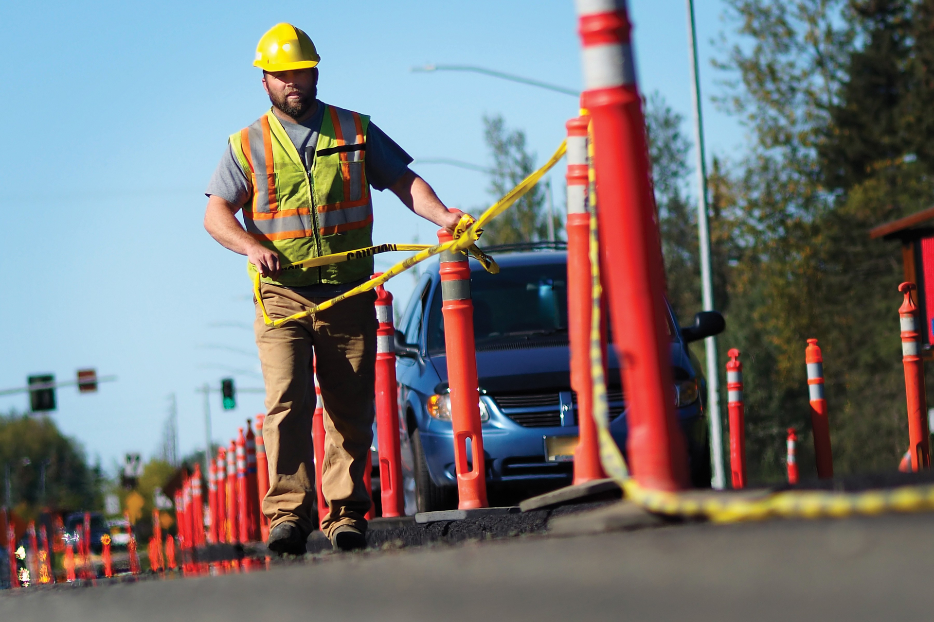 Construction worker in hard hat and safety vest securing yellow caution tape around orange traffic cones.