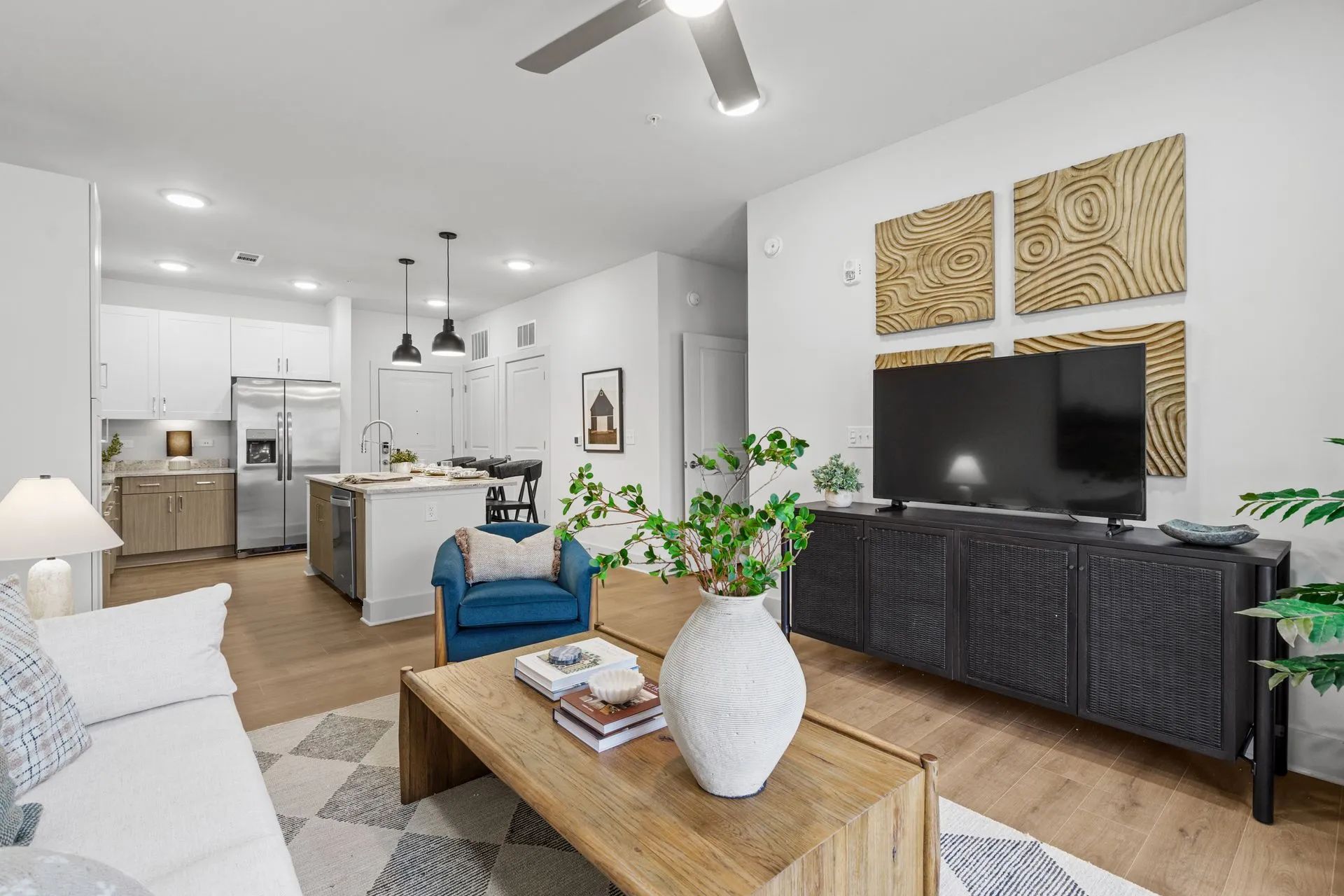 Modern living room with open kitchen. White walls, wood flooring, black TV console, and neutral furniture.