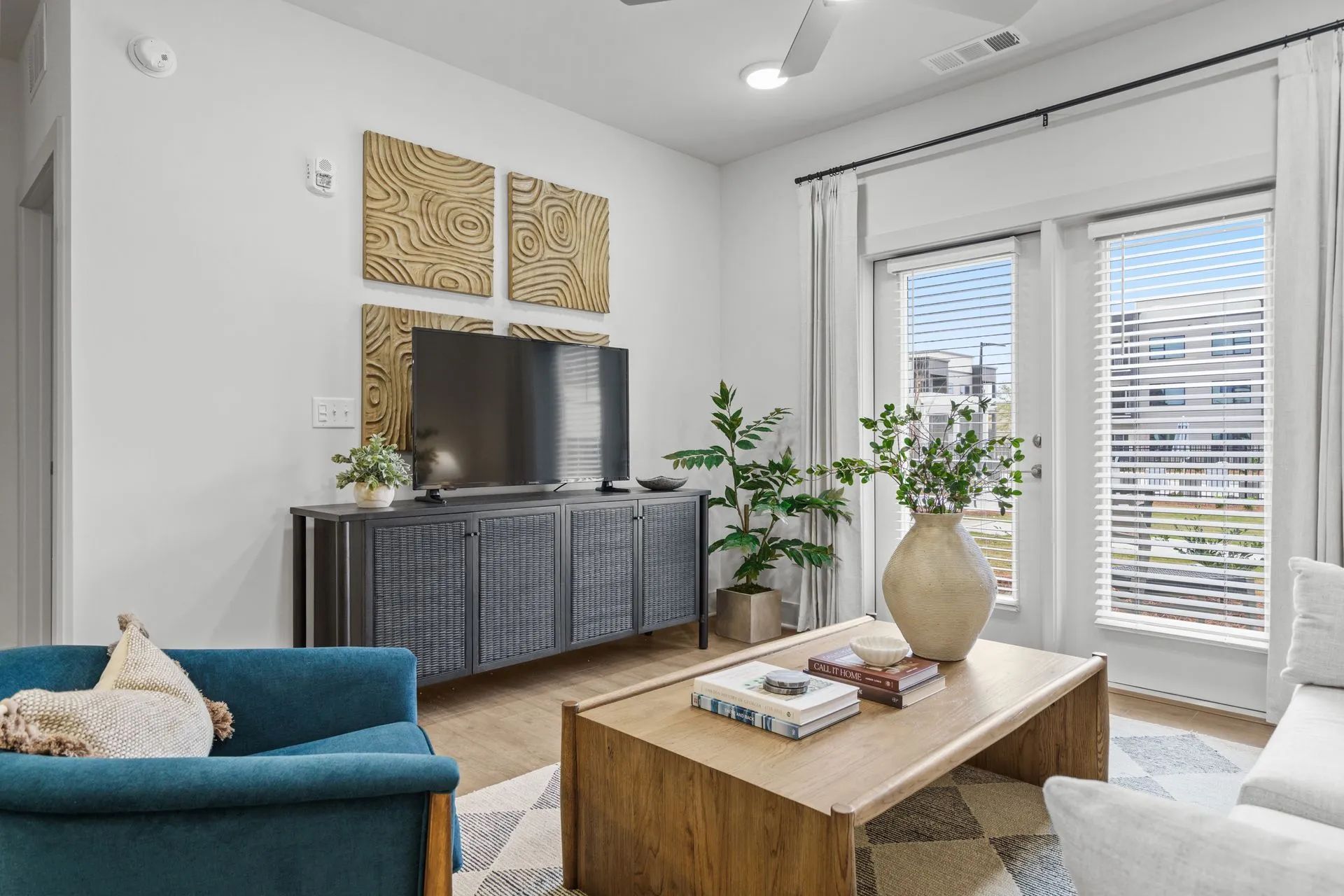 Living room with blue accents, TV, and balcony access. Wooden decor, plants, and beige and white tones.