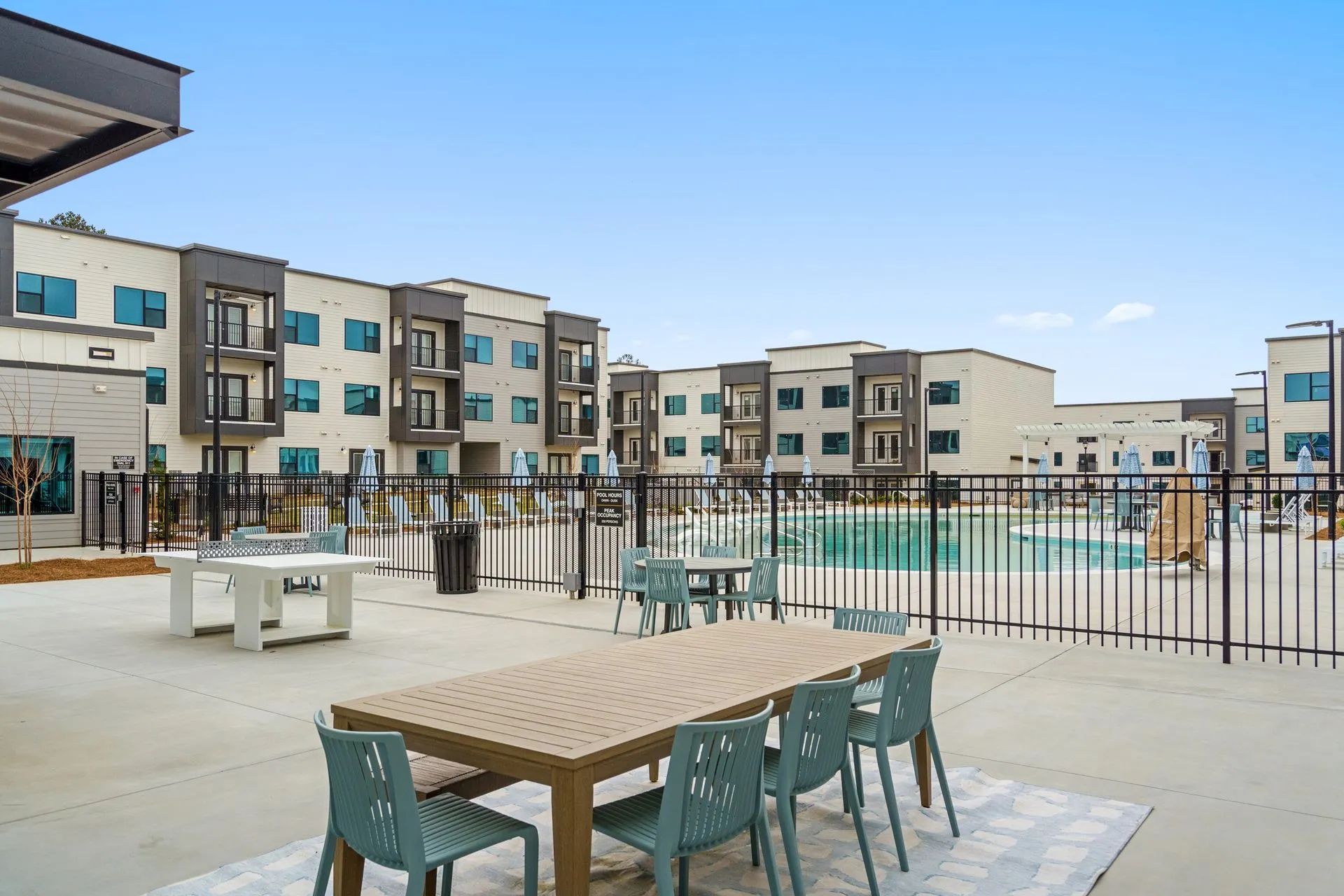 Outdoor patio with tables, chairs, and a swimming pool, in front of modern apartment buildings under a blue sky.