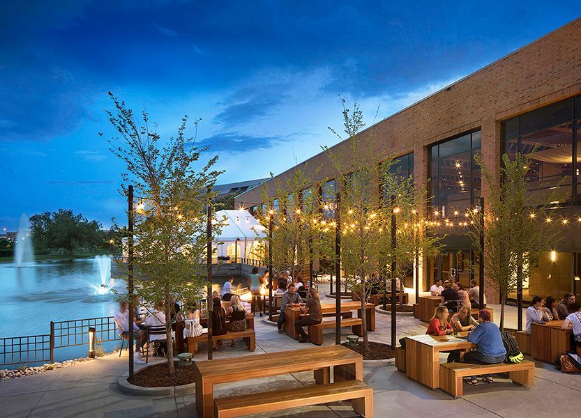 Outdoor restaurant patio at dusk, with diners at wooden tables, a water fountain, and string lights.