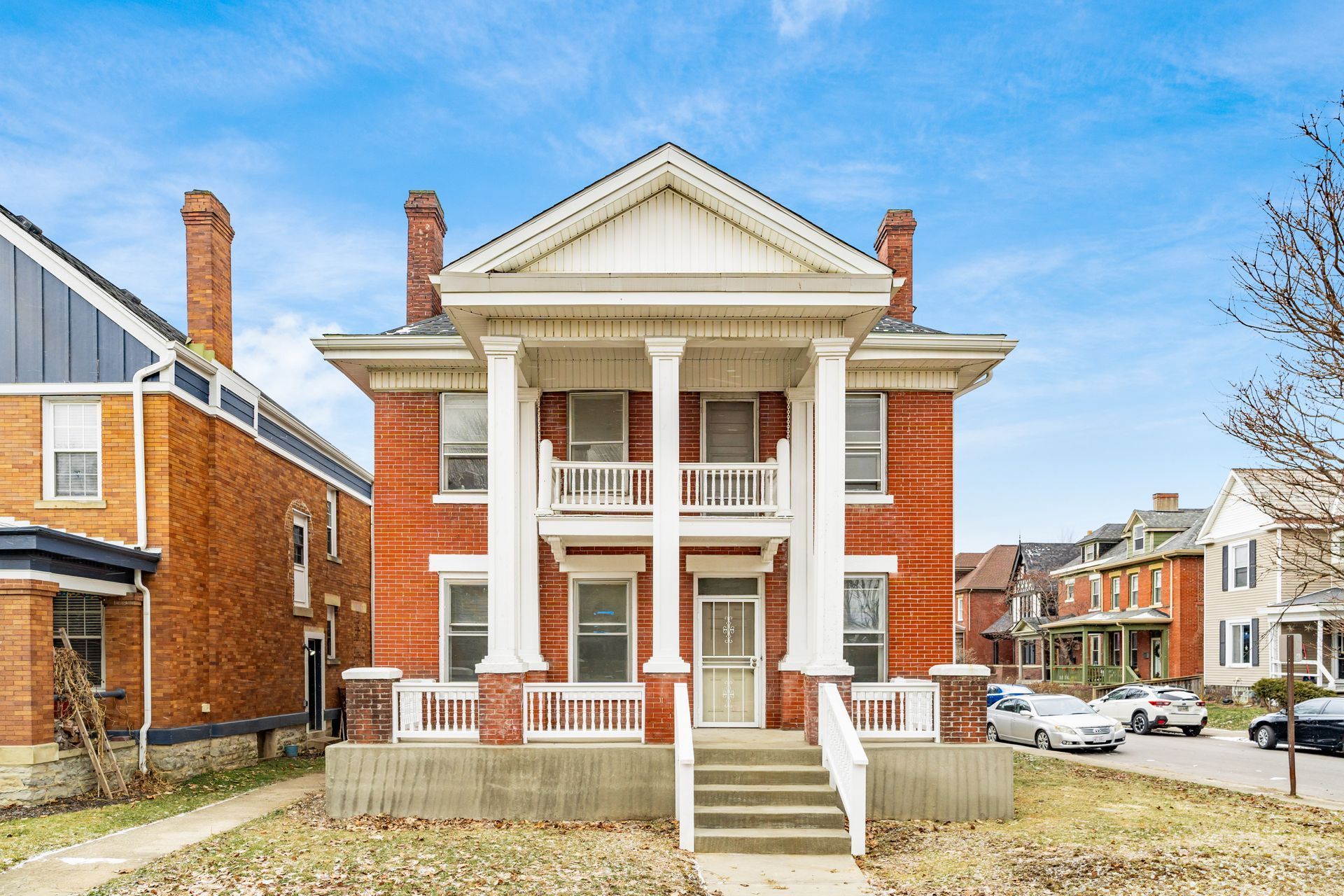 Historic white-columned brick house with front steps, red shutters, and neighboring homes on a sunny street