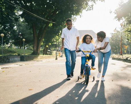 A child rides a blue bike while two adults walk alongside on a sunlit park path.