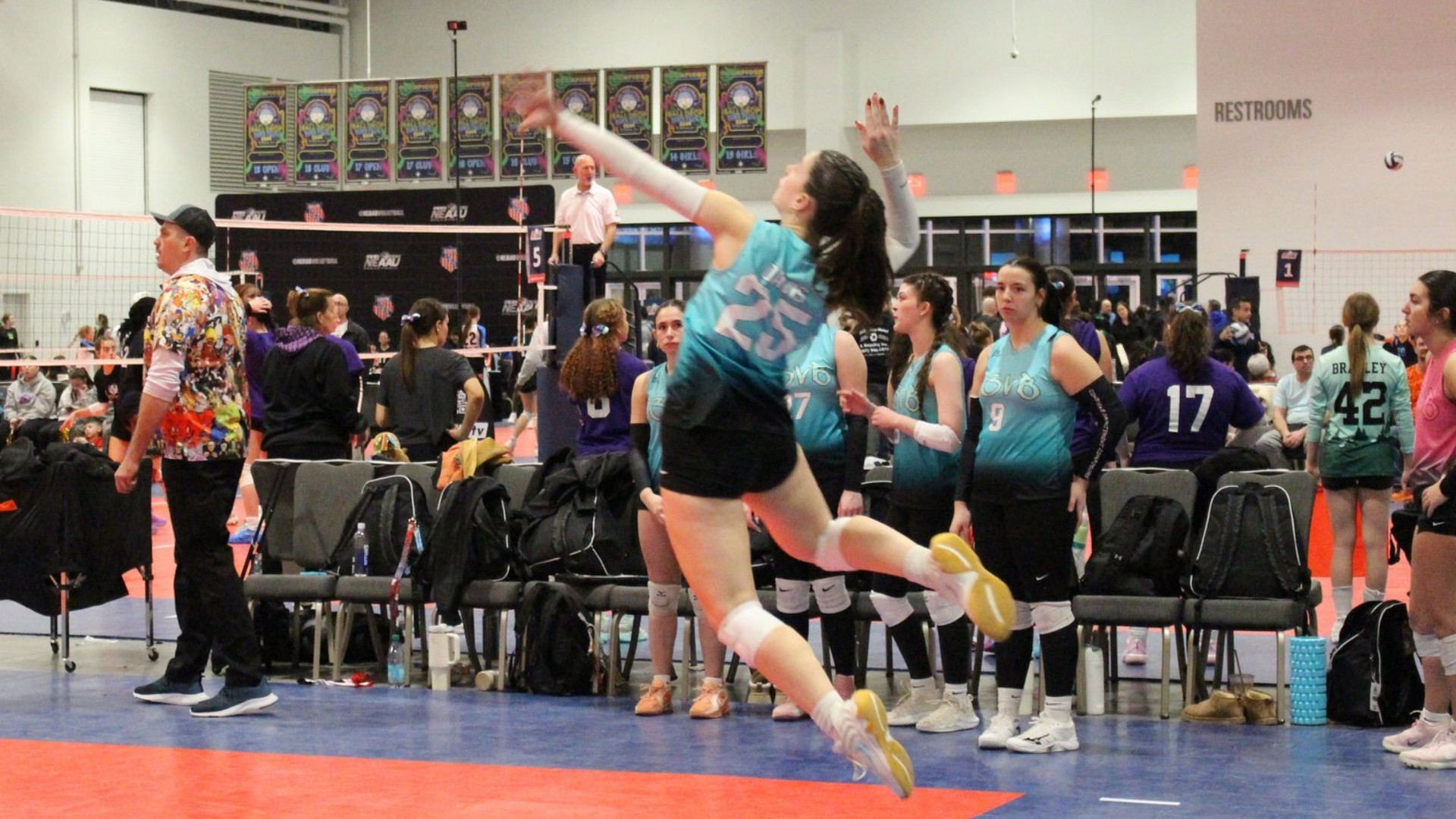 Volleyball player jumping to spike ball during game. Wearing teal jersey. Indoor setting.