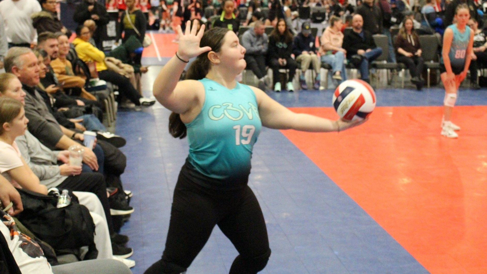 Volleyball player in teal jersey with number 19 about to serve the ball in a crowded gymnasium.